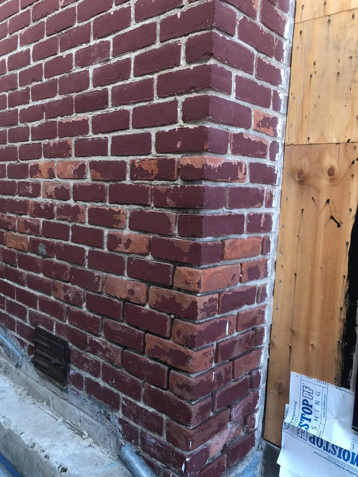 The corner of a red-brown brick chimney where it meets the roof, with metal flashing, a vent and wood sheathing under house wrap beside it
