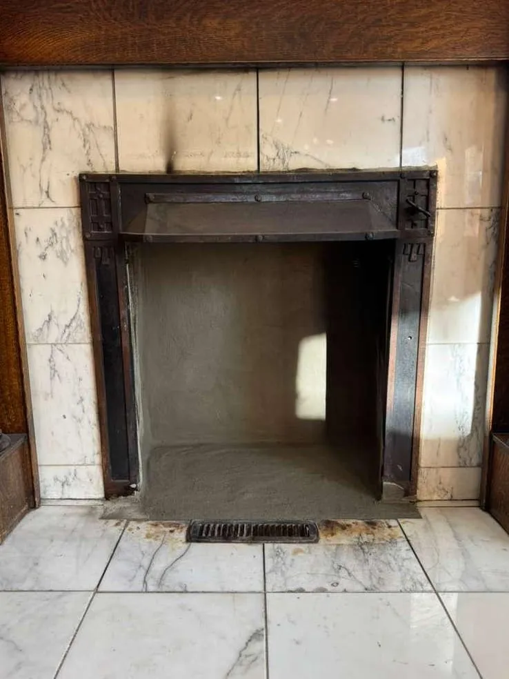 A white marble fireplace with a dark iron frame and a clean, empty firebox with a small ash drain in the hearth