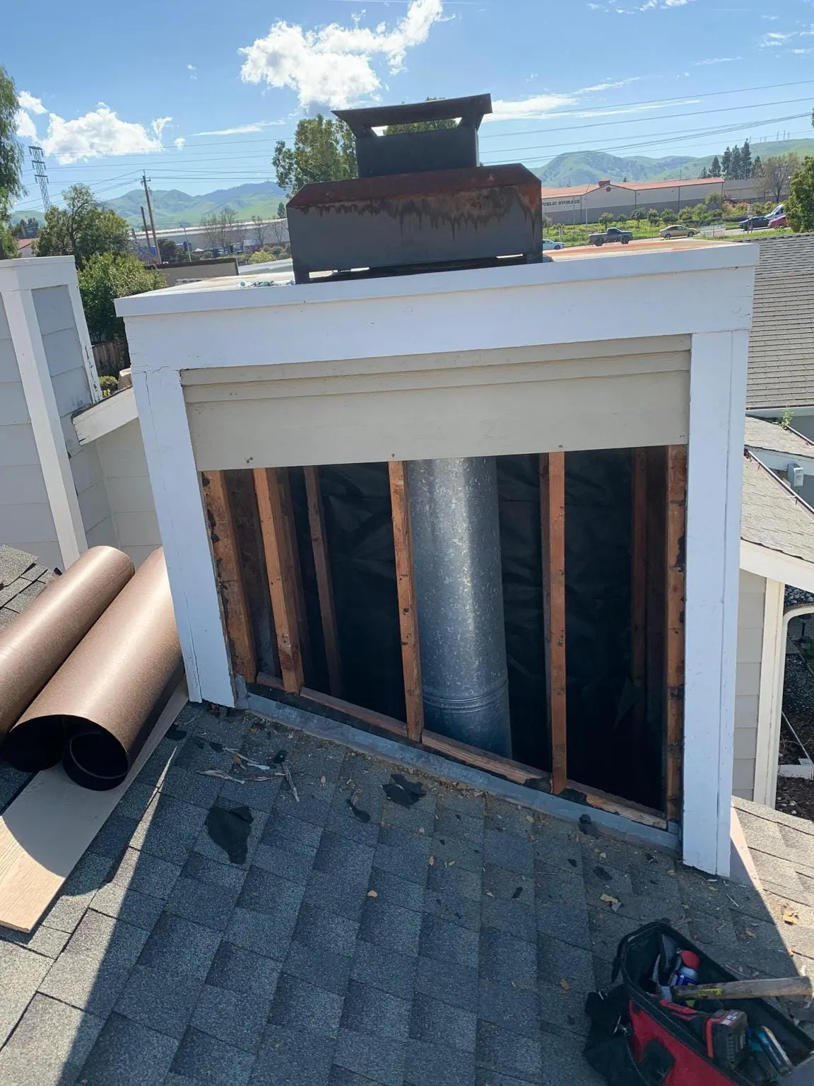 A framed chimney chase opened at the side to show a metal flue pipe inside, on a shingled roof with roofing tools beside it