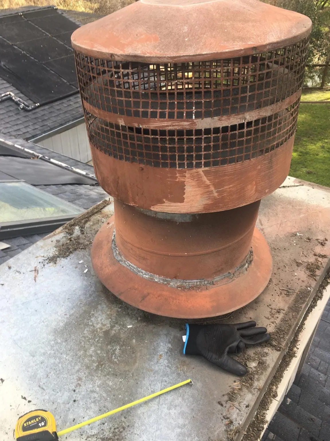 A rust-red metal chimney cap with a wire mesh band, on a metal roof deck, with a work glove and a tape measure beside it