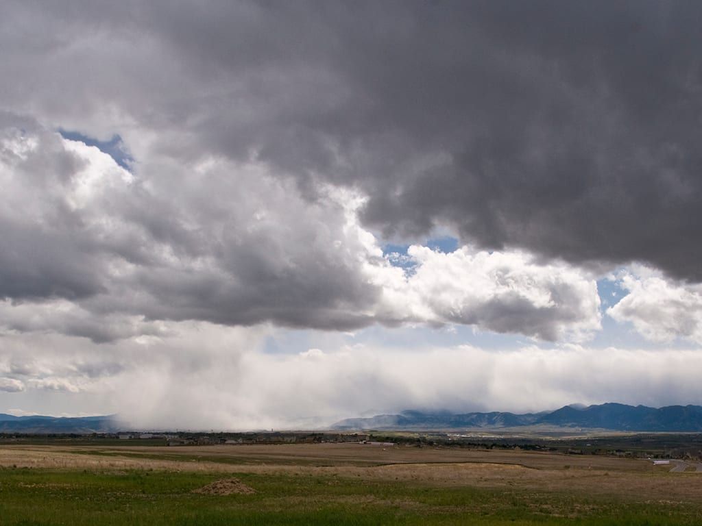 A heavy gray storm cloud over open plains, rain falling in the distance and the Front Range as a thin blue line under it
